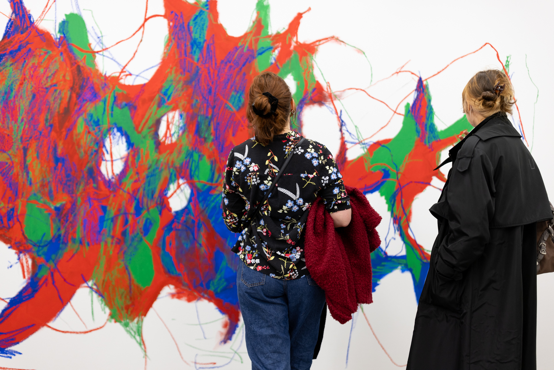 Zwei Menschen, die von hinten zu sehen sind, stehen vor einer bunten Wandmalerei, die sich netzartig in den Farben grün, rot und blau über diese ausbreitet. 