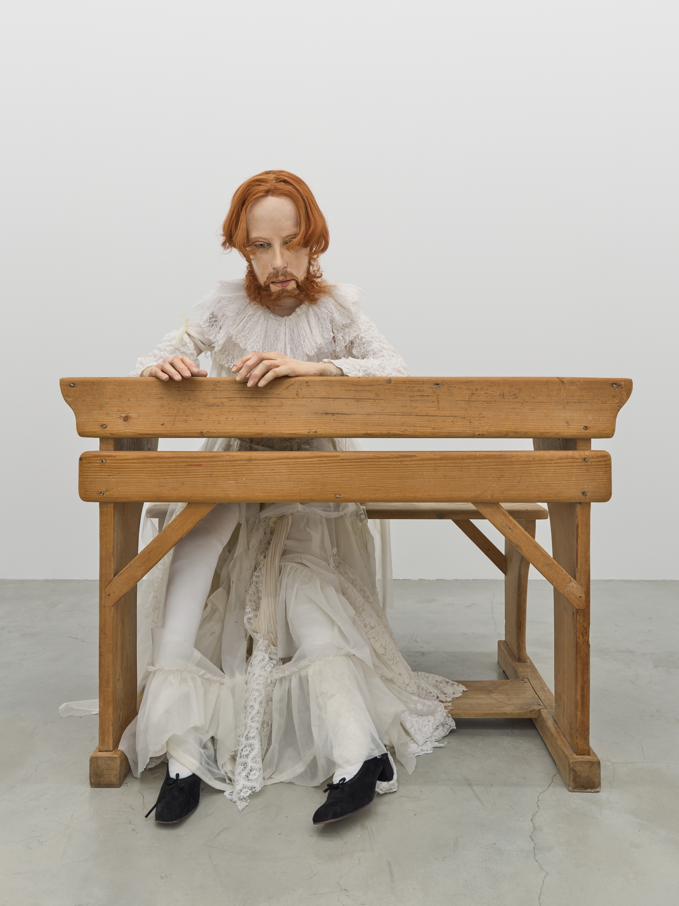 Eine Puppe mit oragenen Haaren, die den Erzherzog Luziwuzi darstellt, sitzt in einem zerissenen, weißen Kleid alleine an einer Schulbank aus dem 20. Jahrhundert.